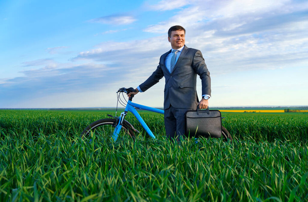 un uomo d'affari in piedi con una bicicletta su un campo di erba verde e guarda in lontananza, vestito in un abito da lavoro, ha una valigetta e documenti, bella natura in primavera, un concetto di business - Foto, immagini