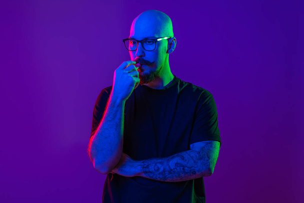 Young bald man using electronic cigarette, vaping in studio. color filters. Vaping concept - 写真・画像