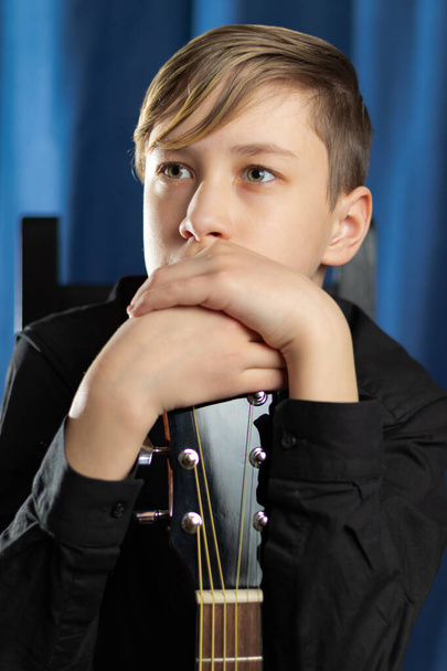 Een tiener in een zwart shirt met een akoestische gitaar op het podium na een concert op een blauwe achtergrond. Hobby. Muziekinstrumenten. Selectieve focus. Portret - Foto, afbeelding