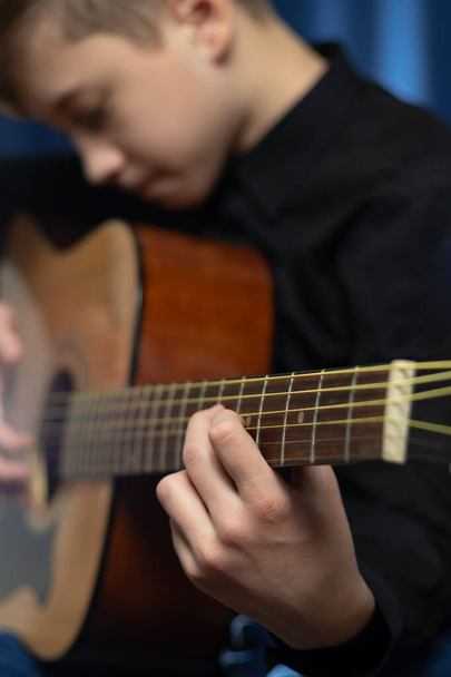 Un adolescente con una camisa negra toca la guitarra acústica en un concierto sobre un fondo azul. Hobby. Instrumentos musicales. Enfoque selectivo. Retrato - Foto, imagen