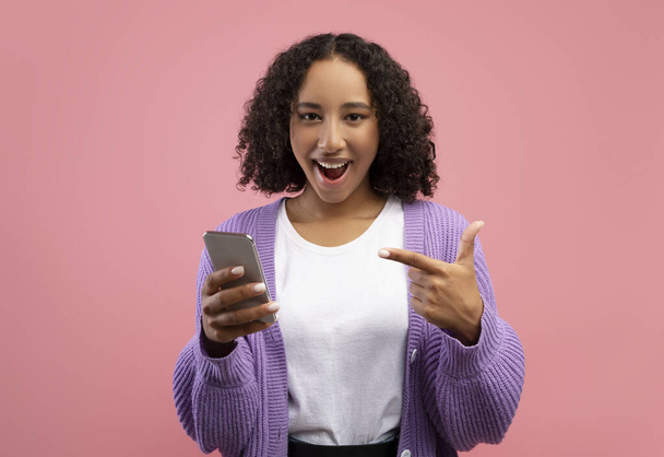 Возбужденная молодая черная женщина, указывающая на смартфон, счастлива из-за огромной онлайн-распродажи на розовом фоне студии - Фото, изображение