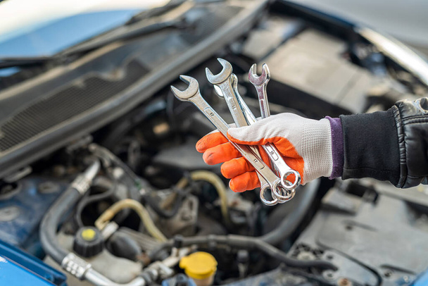 mano del mecánico en un guante especial sostiene un conjunto de herramientas para el coche cerca del coche con una capucha abierta. concepto de reparación de coches - Foto, Imagen