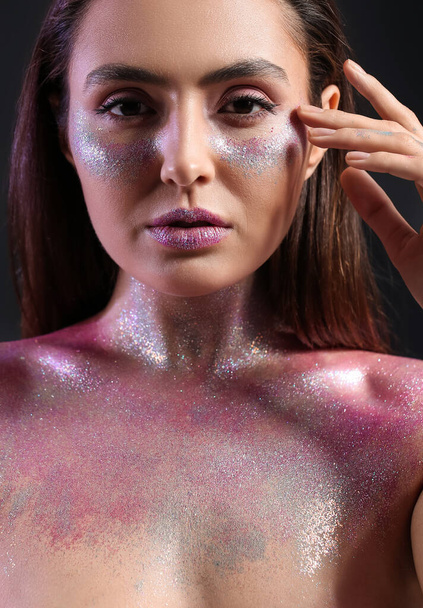 Beautiful young woman with glitters on her face and body against dark background - Photo, Image