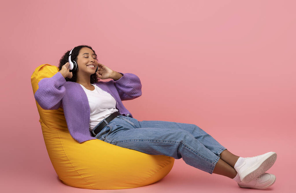 Pełna długość spokojnej młodej czarnej kobiety słuchającej muzyki w słuchawkach, odpoczywającej w fotelu worek fasoli na różowym tle - Zdjęcie, obraz