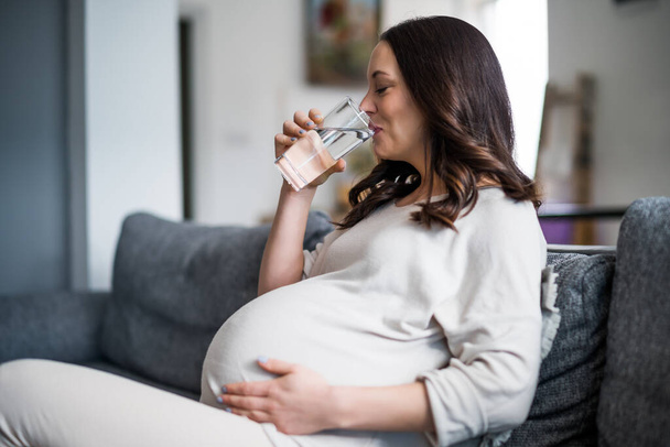 Pregnant woman relaxing at home. She is sitting on bed and drinking water. - Foto, Imagem