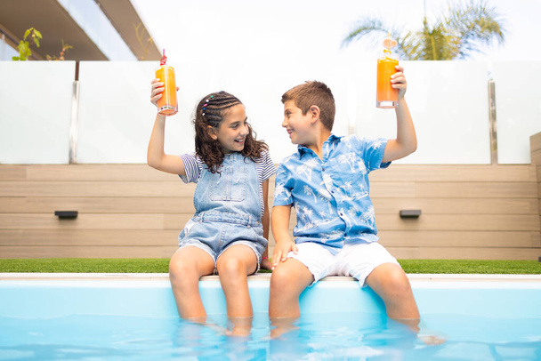 Dvě vtipné děti pronášejí přípitek s pomerančovým džusem v bazénu - Fotografie, Obrázek