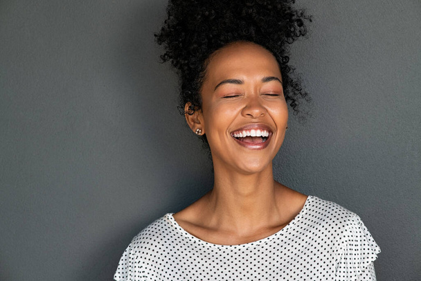 Portrait of cheerful young woman laughing with closed eyes against grey background. Happy african american girl with curly hair smiling against gray wall. Close up face of african student smiling with copy space. - 写真・画像