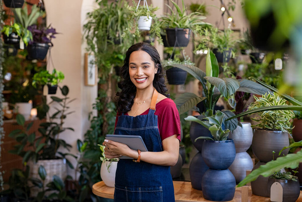 Portrait of smiling young latin woman florist standing and holding digital tablet at floral shop. Successful flower shop owner standing in plant store wearing apron and looking at camera. Happy mixed race entrepreneur in flower shop surrounded by gre - Фото, изображение