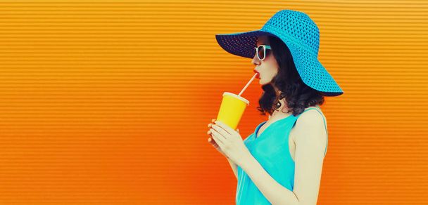 Portrait of beautiful young woman drinking fresh juice from cup wearing summer hat on orange background, blank copy space for advertising text - Foto, Imagem