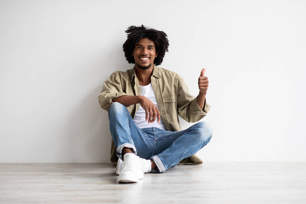Vrolijke zwarte Millennial Guy zit op de vloer en toont duim omhoog - Foto, afbeelding