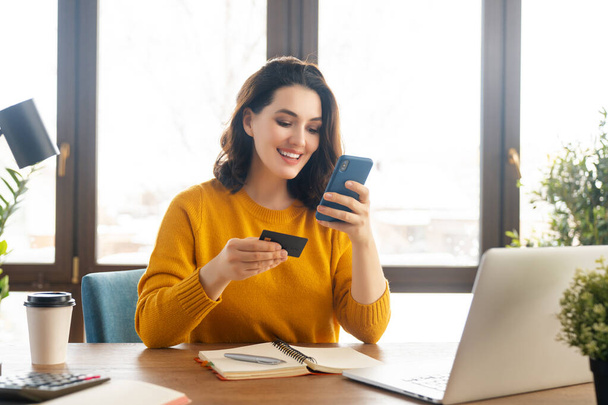 Woman is doing online purchases. Girl is doing shopping using phone and credit card at home. - Foto, imagen