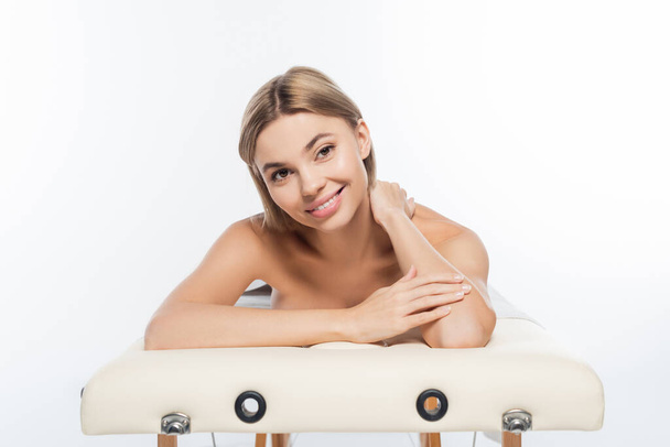 mujer rubia positiva con los hombros desnudos acostado en la mesa de masaje en blanco - Foto, Imagen