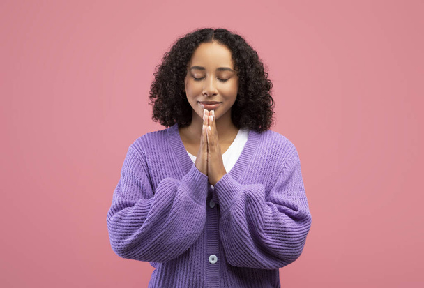 Békés fiatal afro-amerikai nő imádkozik Istenhez csukott kézzel és csukott szemmel rózsaszín stúdió háttér - Fotó, kép