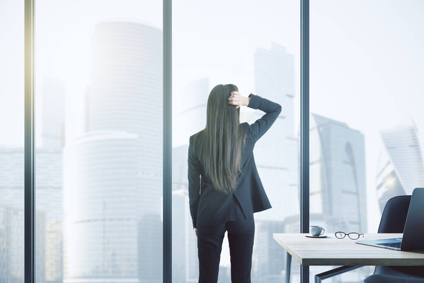 Back view of thoughtful young businesswoman standing in modern office with workplace desktop and panoramic city view. Future and success concept - Foto, Bild