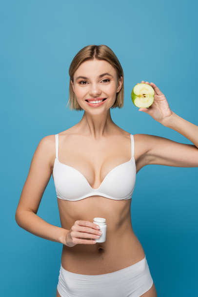 cheerful woman in white lingerie holding bottle with vitamins and apple half isolated on blue  - 写真・画像