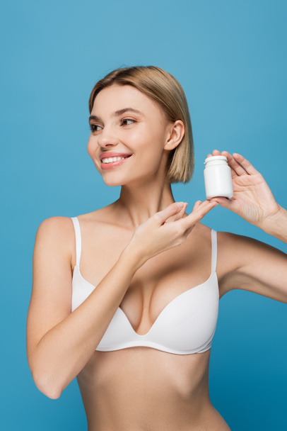 happy young woman in white bra holding bottle with vitamins isolated on blue  - Foto, imagen