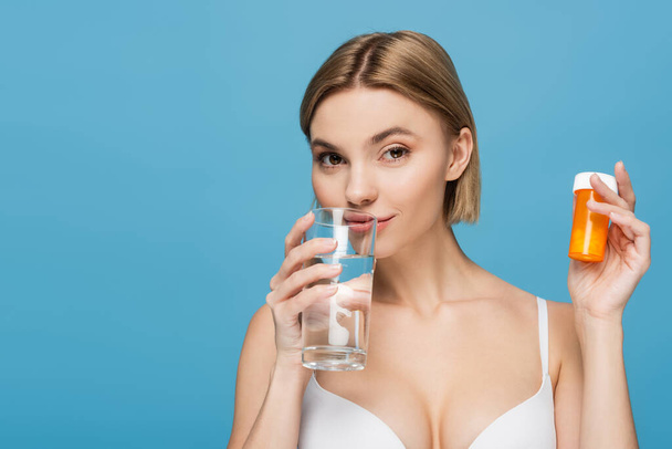 mujer joven en sujetador blanco sosteniendo botella con vitaminas y vaso de agua aislado en azul  - Foto, Imagen