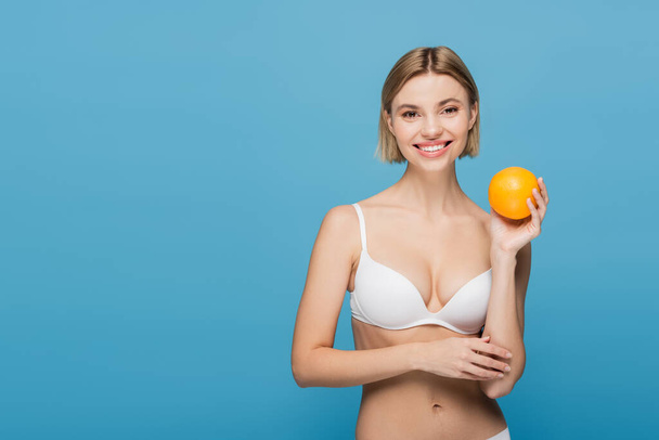 усміхнена молода жінка в білому бюстгальтері тримає стиглий апельсин ізольовано на синьому
  - Фото, зображення