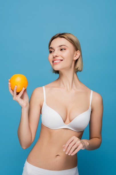 cheerful young woman in white bra holding ripe orange isolated on blue  - Фото, изображение