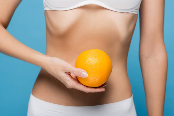 cropped view of young woman in white bra holding ripe orange isolated on blue  - Valokuva, kuva