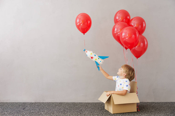 Happy child playing with toy rocket at home. Kid pretend to be astronaut. Boy sitting in cardboard box with red balloons. Imagination and children dream concept - Foto, Bild