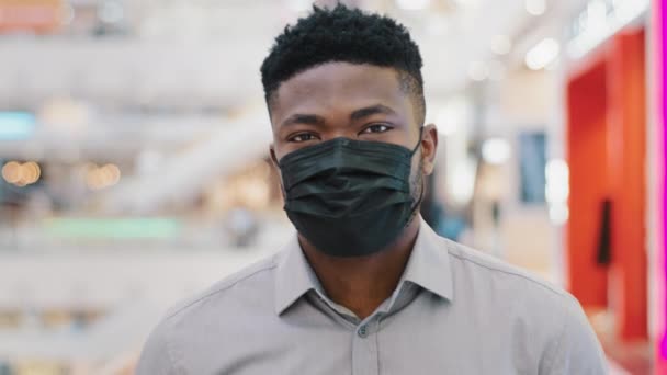Nahaufnahme junger afrikanisch-amerikanischer Mann in medizinischer Schutzmaske, der in einem Einkaufszentrum spazieren geht und die Kamera anschaut, wie das covid19-Virus während einer Pandemie vor Bakterien geschützt wird - Filmmaterial, Video