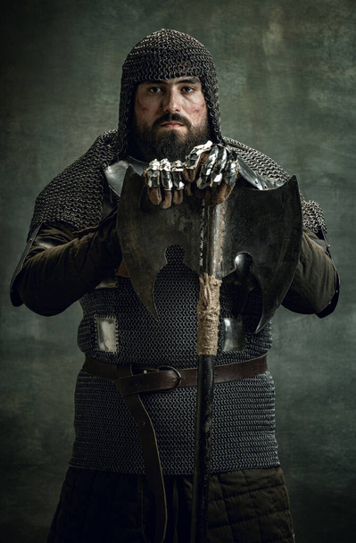 Vintage portrait of brutal seriuos man, medieval warrior or knight with wounded face in chain mail holding ax isolated over dark background. Comparison of eras - Foto, immagini