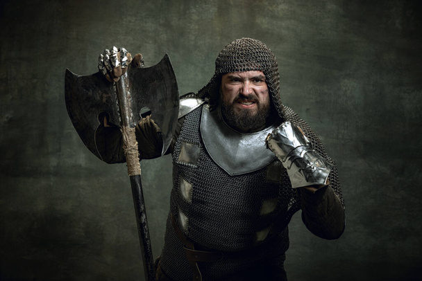 Vintage portrait of brutal seriuos man, medieval warrior or knight with wounded face in chain mail holding ax isolated over dark background. Comparison of eras - Foto, imagen