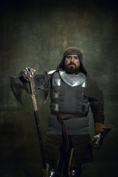 Vintage portrait of brutal seriuos man, medieval warrior or knight with wounded face in chain mail holding ax isolated over dark background. Comparison of eras - Фото, изображение