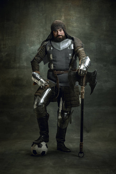 Vintage portrait of brutal seriuos man, medieval warrior or knight with wounded face in chain mail holding ax isolated over dark background. Comparison of eras - Φωτογραφία, εικόνα