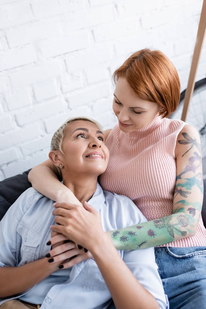 tattooed woman hugging happy lesbian girlfriend while looking at each other at home  - Photo, Image