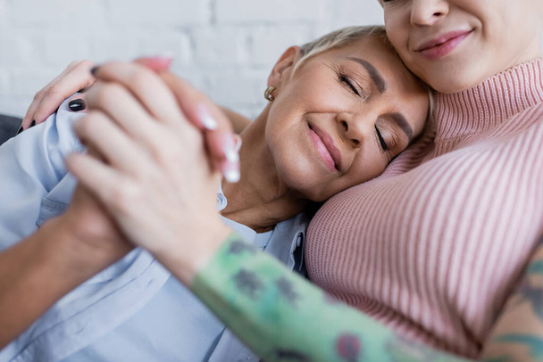 happy lesbian woman with closed eyes holding hands with blurred girlfriend at home - Фото, зображення