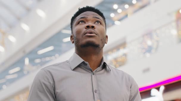 Close-up male portrait young african american man student tourist standing in huge shopping and entertainment center looks around in amazement stunned by scale building sincerely surprised admires - Footage, Video