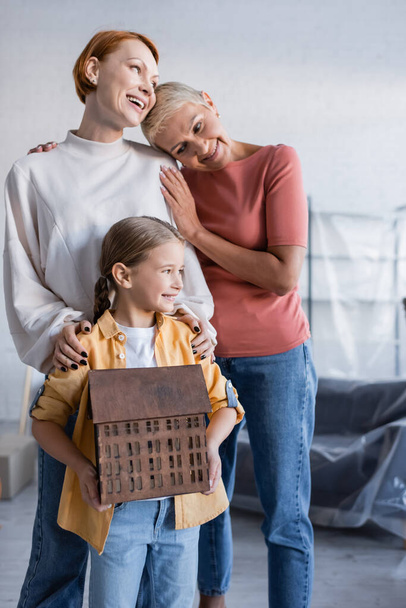 preteen girl holding house model near pleased same sex parents in new home - Photo, Image