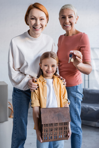 lesbian couple looking at camera near happy adopted daughter holding house model - Photo, Image
