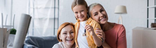 cheerful lesbian women looking at camera near adopted daughter holding key from new apartment, banner - Photo, Image