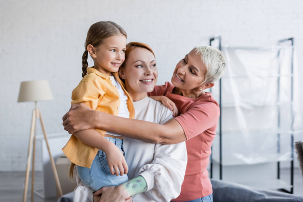 cheerful lesbian woman embracing girlfriend with adopted daughter in new home - Foto, Imagem