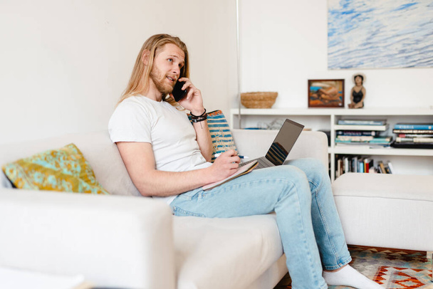 Bearded blonde man talking on cellphone while working with laptop at home - 写真・画像