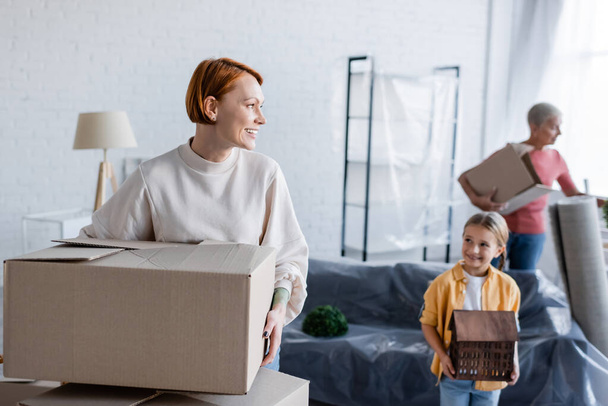 smiling woman holding carton package near adopted daughter with house model - Photo, Image
