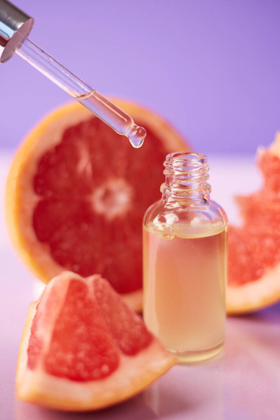 Ätherisches Zitrus- oder Grapefruitöl in Glasflasche mit frischer Grapefruit auf trendigem lila Hintergrund. Kur- und Pflegeprodukt - Foto, Bild