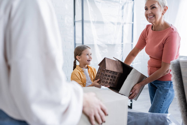 cheerful child holding house model near same sex parents with carton boxes - Fotó, kép