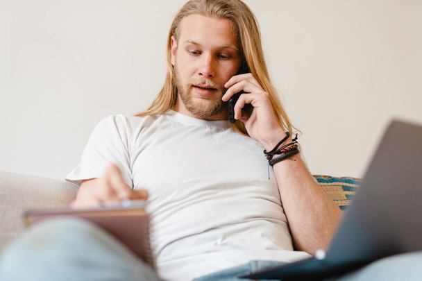 Bearded blonde man talking on cellphone while working with laptop at home - Foto, immagini