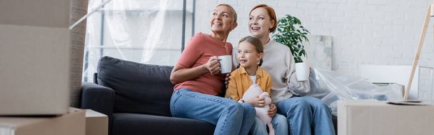szczęśliwy lesbijki i adoptowane córka siedzi na kanapie w nowym mieszkaniu i patrząc daleko, sztandar - Zdjęcie, obraz