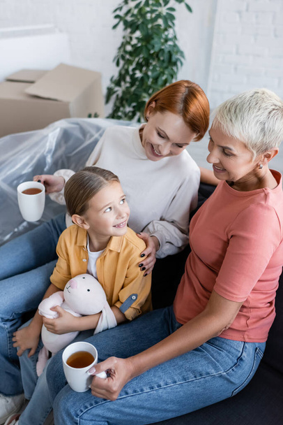 mulheres lésbicas com xícaras de chá e menina com coelho de brinquedo sorrindo no sofá no novo apartamento - Foto, Imagem