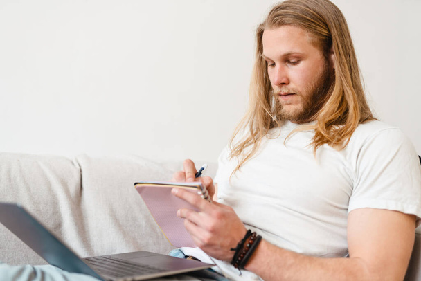 Bearded blonde man writing down notes while sitting on couch at home - Foto, Bild