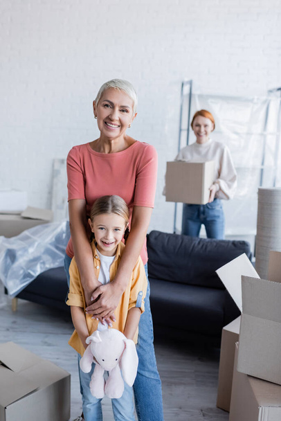 happy lesbian woman with adopted daughter smiling at camera near carton boxes in new home - Foto, imagen
