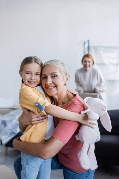 alegre lesbiana mujer abrazando adoptivo hija y mirando cámara en nuevo hogar - Foto, Imagen