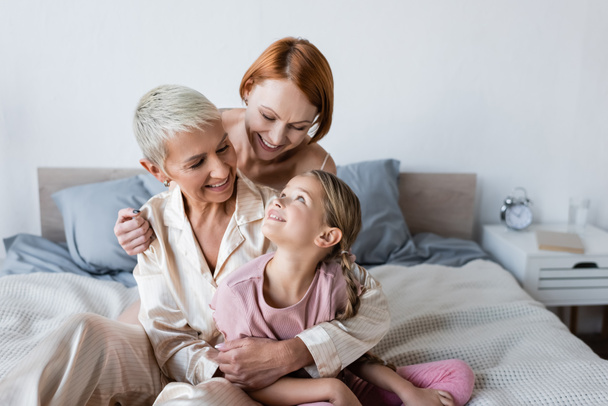 Woman hugging girlfriend in pajamas and child on bed  - Photo, Image