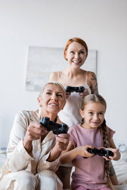KYIV, UKRAINE - 2021年12月8日:パジャマの女性がベッドルームでガールフレンドの近くの娘とビデオゲームをプレイ  - 写真・画像