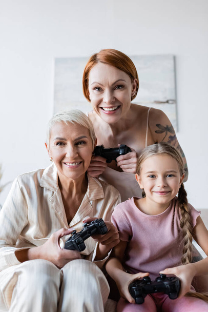 KYIV, UKRAINE - DECEMBER 8, 2021: Happy lesbian couple and adopted daughter holding joysticks and looking at camera at home  - Foto, Bild